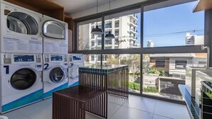 Uma lavanderia com várias máquinas de lavar, um cesto de madeira e uma janela grande com vista para edifícios.
