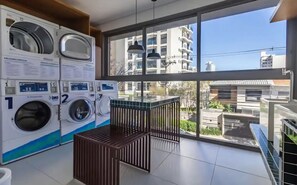 A laundry room with multiple washing machines, a wooden crate, and a large window with a view of buildings. - Queen bed with Emma mattress, air conditioning, double shower, and Nespresso coffee machine. (São Paulo)