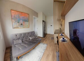 A modern living room with a grey sofa, a painting, a wooden dining table with blue and white plates, and a TV. - Queen bed with Emma mattress, air conditioning, double shower, and Nespresso coffee machine. (São Paulo)