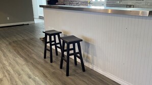 A bar area with wooden flooring, two stools, and a counter with a light fixture above.
