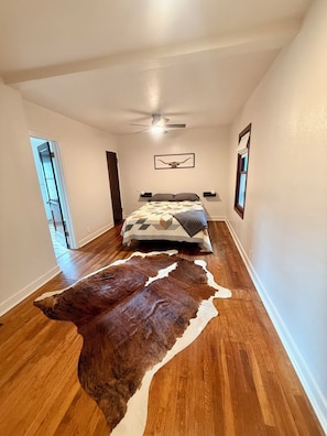 Quarto com cama, ventilador de teto, tapete de couro de vaca e porta que dá para outro cômodo.