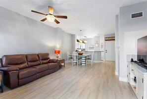 Una sala de estar moderna con un sofá de cuero marrón, un comedor con mesa y sillas, una cocina con un refrigerador blanco y un ventilador de techo.
