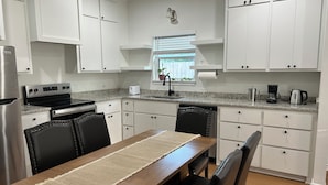 Una cocina moderna con armarios blancos, encimera de granito, electrodomésticos de acero inoxidable y una zona de comedor con mesa y sillas de madera.