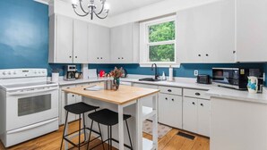 A modern kitchen with white cabinets, a wooden table, black bar stools, a chandelier, and a window with a view of greenery. - Gaspereau Heritage House - Annapolis Valley (Wolfville)
