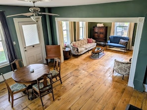 A room with a wooden floor, a dining table with chairs, a sofa, a patterned armchair, a cabinet, and a ceiling fan.