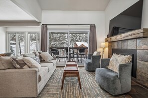 A living room with a sofa, armchair, and a small wooden table. There is a fireplace and a large window with curtains. - 4 O'Clock Lodge D27: Remodeled condo close to town and the slopes - New Listing. (Breckenridge)