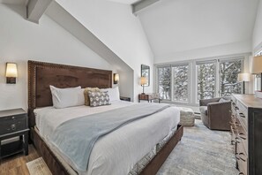 Quarto com uma cama grande, abajures, cômoda e vista para árvores através de amplas janelas.