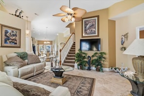 A spacious living area with a ceiling fan, a staircase, a flat-screen TV, and various decorative items. - THE BEACH IS CALLING YOU -Truly One of a Kind Enjoy the Pool, Beach, Golf Cart! (North Myrtle Beach)
