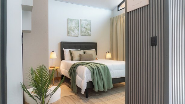 Un dormitorio con cama, dos lámparas de noche, una manta verde, una planta en maceta y dos obras de arte en la pared.