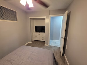 Un dormitorio con cama, televisor sobre un aparador blanco, ventilador de techo y puerta que da a un balcón.
