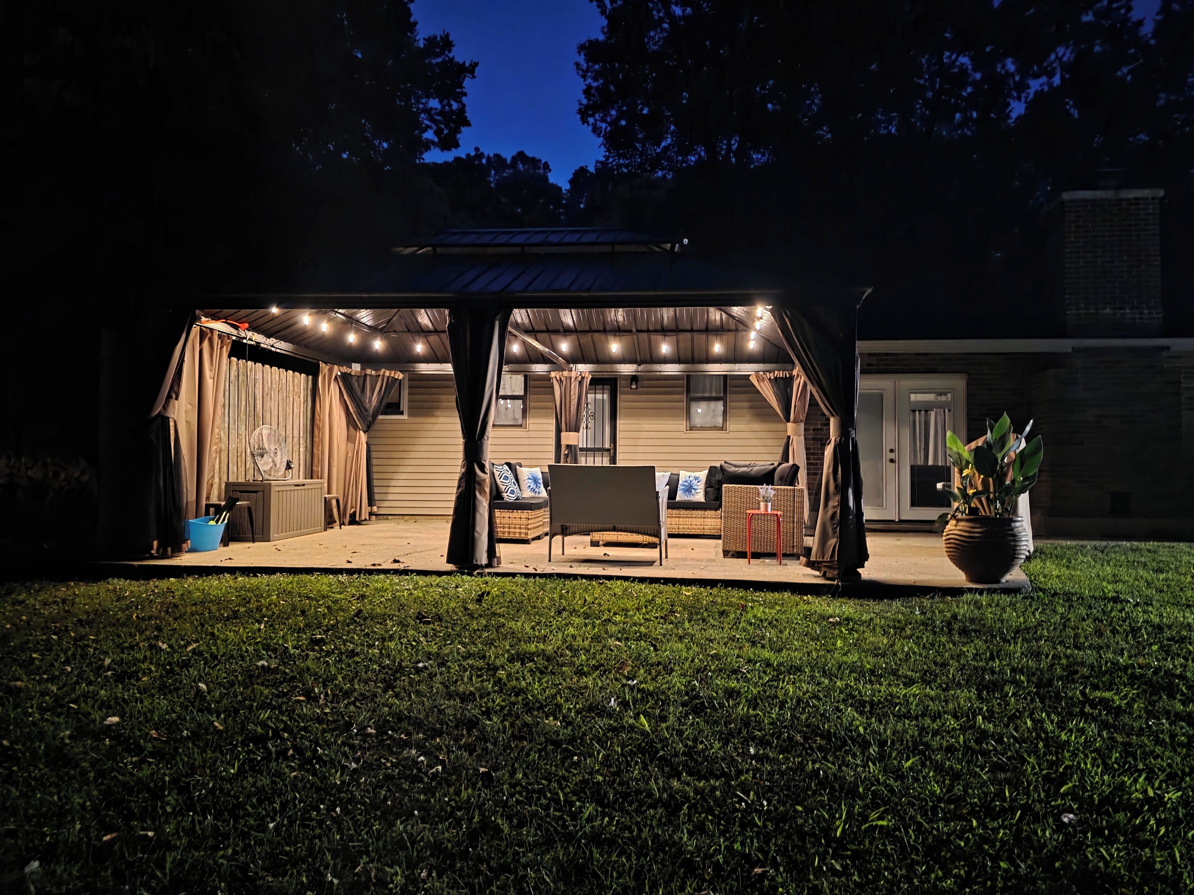 An outdoor covered patio area with a dining table and chairs, a wicker sofa, and a potted plant.