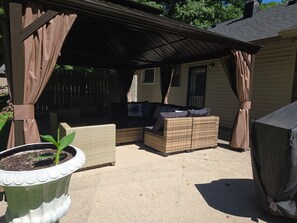 A covered patio area with a wicker sofa, a potted plant, and a grill.