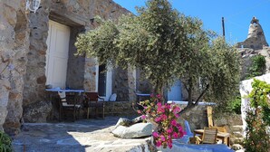 A stone building with a white door, a small table with chairs, and a tree with pink flowers. - Room-charming Studio Rental in Mikonos w (Mykonos)