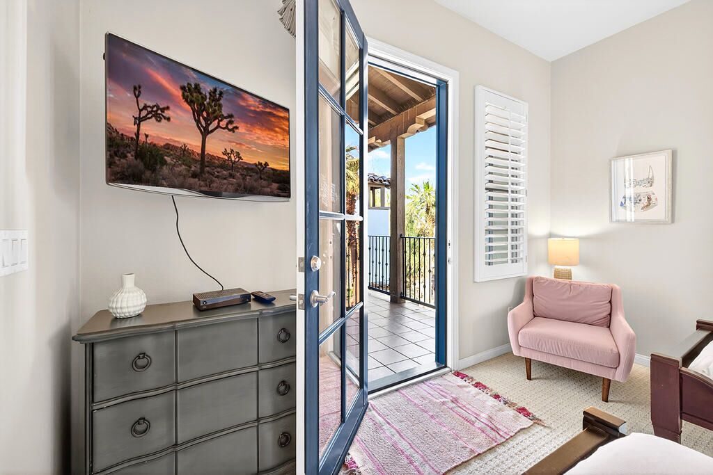 Une pièce comprenant un lit, une commode, un fauteuil rose, un téléviseur et une porte donnant sur un balcon extérieur.