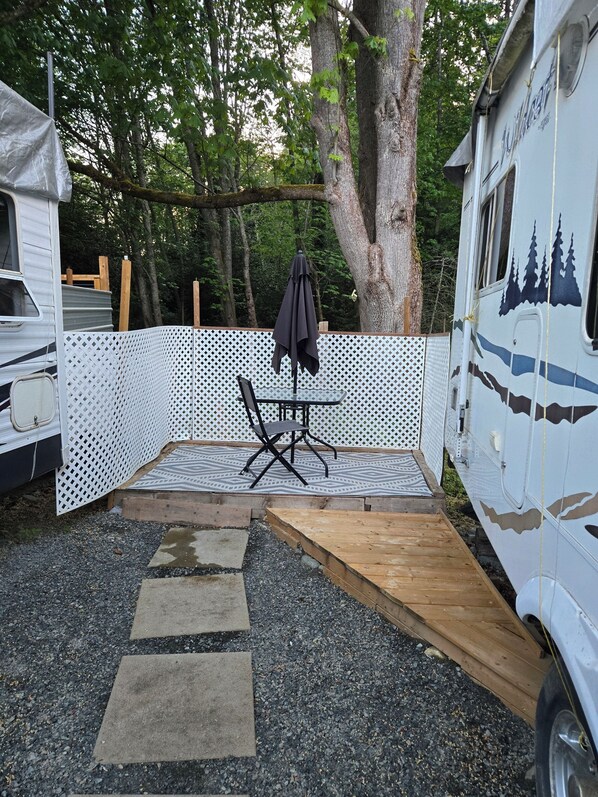 Un terrain de camping équipé d’une table et de chaises, d’un parasol et d’une rampe en bois menant à un véhicule.