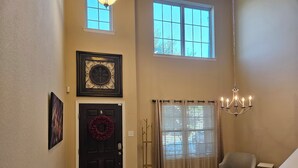 A spacious entryway with a dark door, a wreath, a chandelier, and a large window with blinds. - Peaceful Retreat for Travel Nurses & Corporate Stays (Spring)