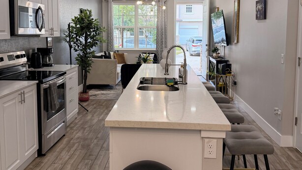 A modern kitchen with white cabinetry, a central island, and a dining area with a view. - Super Calm-Sleeps 13 w Designs & close to Parks! (Kissimmee)