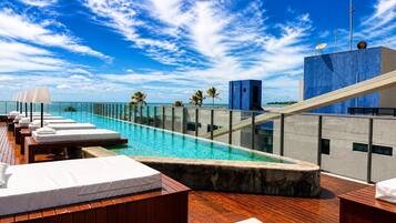 A rooftop infinity pool with lounge chairs, a clear view of the ocean, and a modern building with a blue facade.