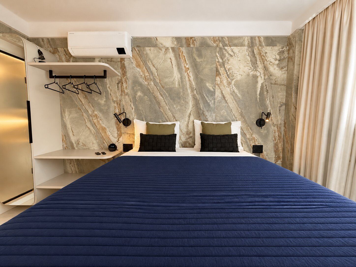 Ein modernes Hotelzimmer mit einem großen Bett, Wänden mit Marmormusterung, einer eingebauten Klimaanlage und einem Kleiderschrank mit Spiegel.