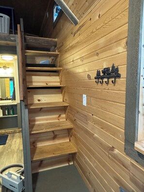 A wooden staircase with a metal handrail, a wall-mounted coat rack, and a power outlet. - Cozy Island Park Tiny Home (Island Park)