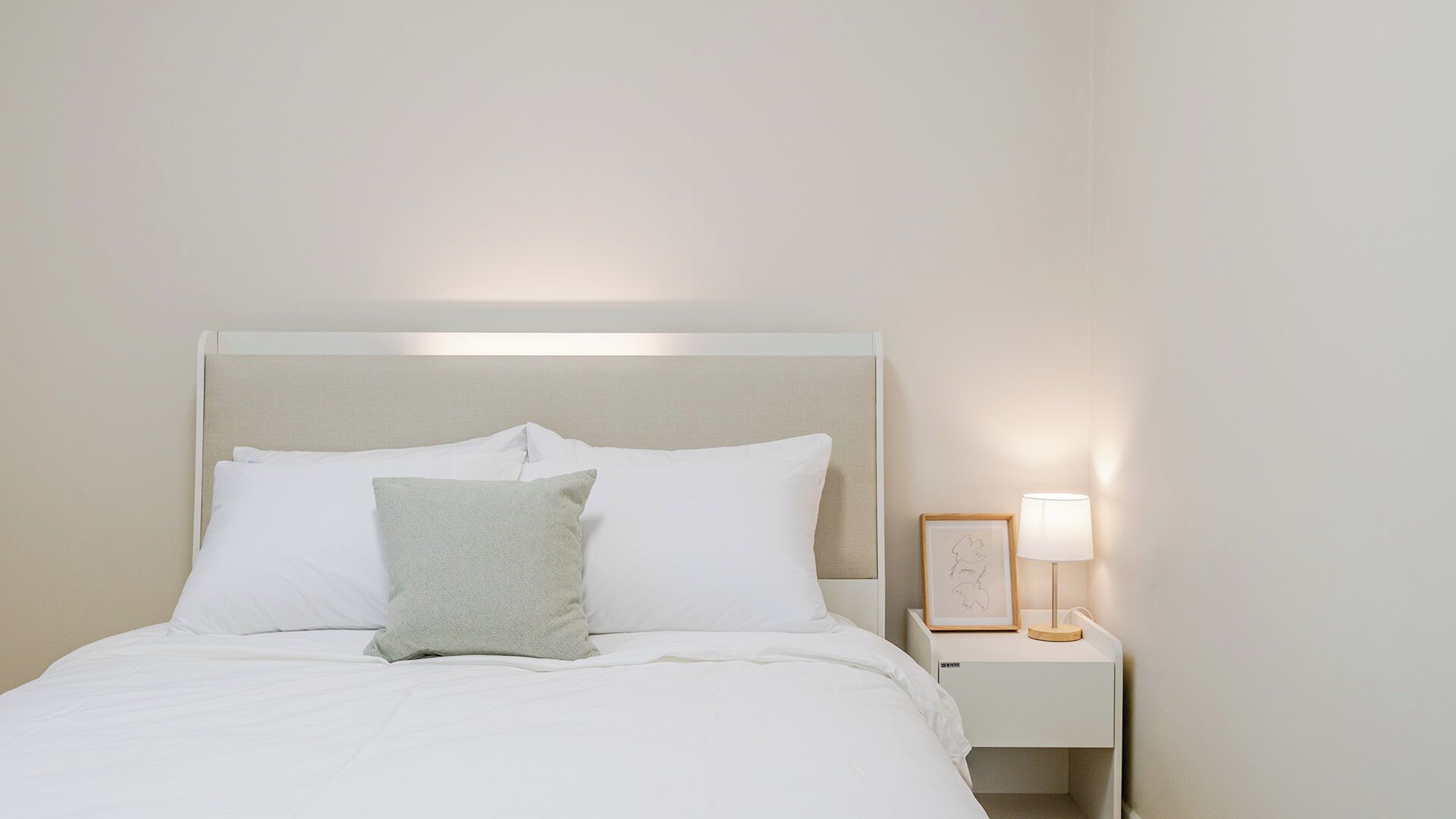 Cama bem arrumada com roupa de cama branca e uma única almofada, mesa de cabeceira com abajur e uma moldura, e uma parede lisa.