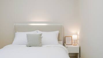 Cama bem arrumada com roupa de cama branca e uma única almofada, mesa de cabeceira com abajur e uma moldura, e uma parede lisa.