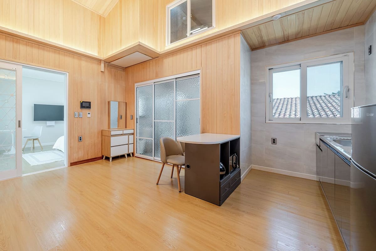 A spacious room with wooden floors, a dining area, a kitchen with a refrigerator, and large windows.