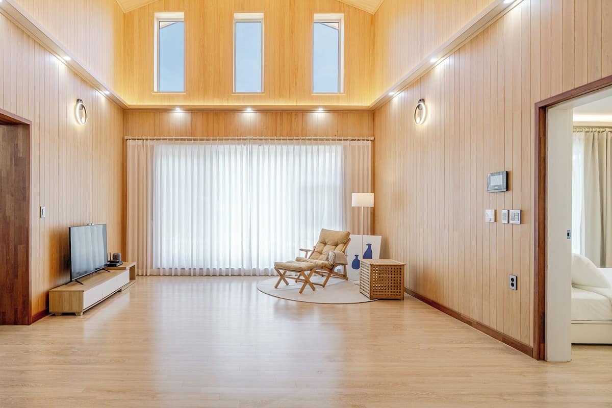 A spacious living area with a high ceiling, wooden walls, a large window with curtains, a flat-screen TV, a sofa, and a refrigerator.