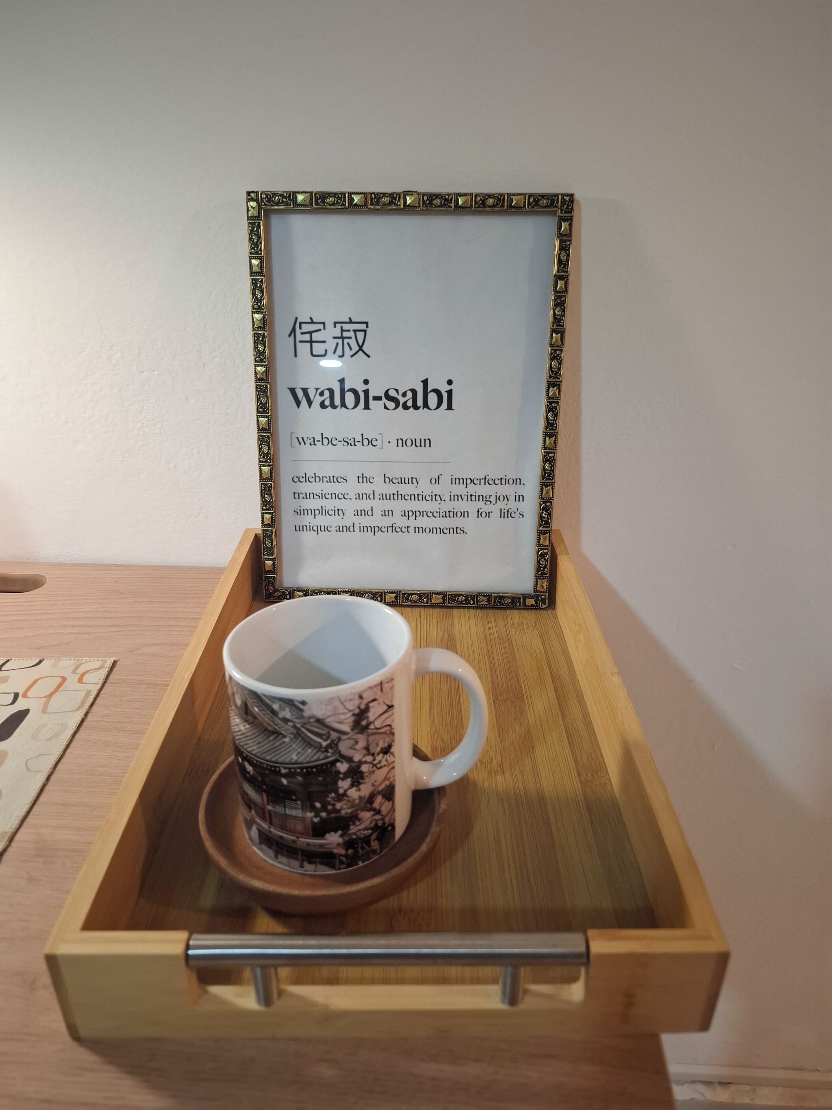 A framed sign with the word 'wabi-sabi' and its definition, placed on a wooden tray with a cup.