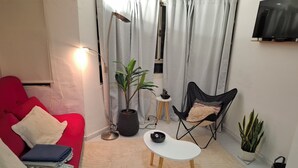 Moderni olohuone, jossa on punainen sohva, pieni valkoinen pöytä, musta tuoli, lattiavalaisin, ruukkukasvi ja seinälle asennettu televisio.