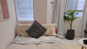 Sängyssä on harmaa peitto, tumma tyyny ja beigenvärinen viltti. Siellä on myös ruukussa oleva kasvi ja pieni pöytä, jonka päällä on kulho.