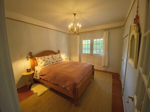 Een slaapkamer met een houten bed, een nachtkastje, een lamp, een raam met gordijnen en een kroonluchter.
