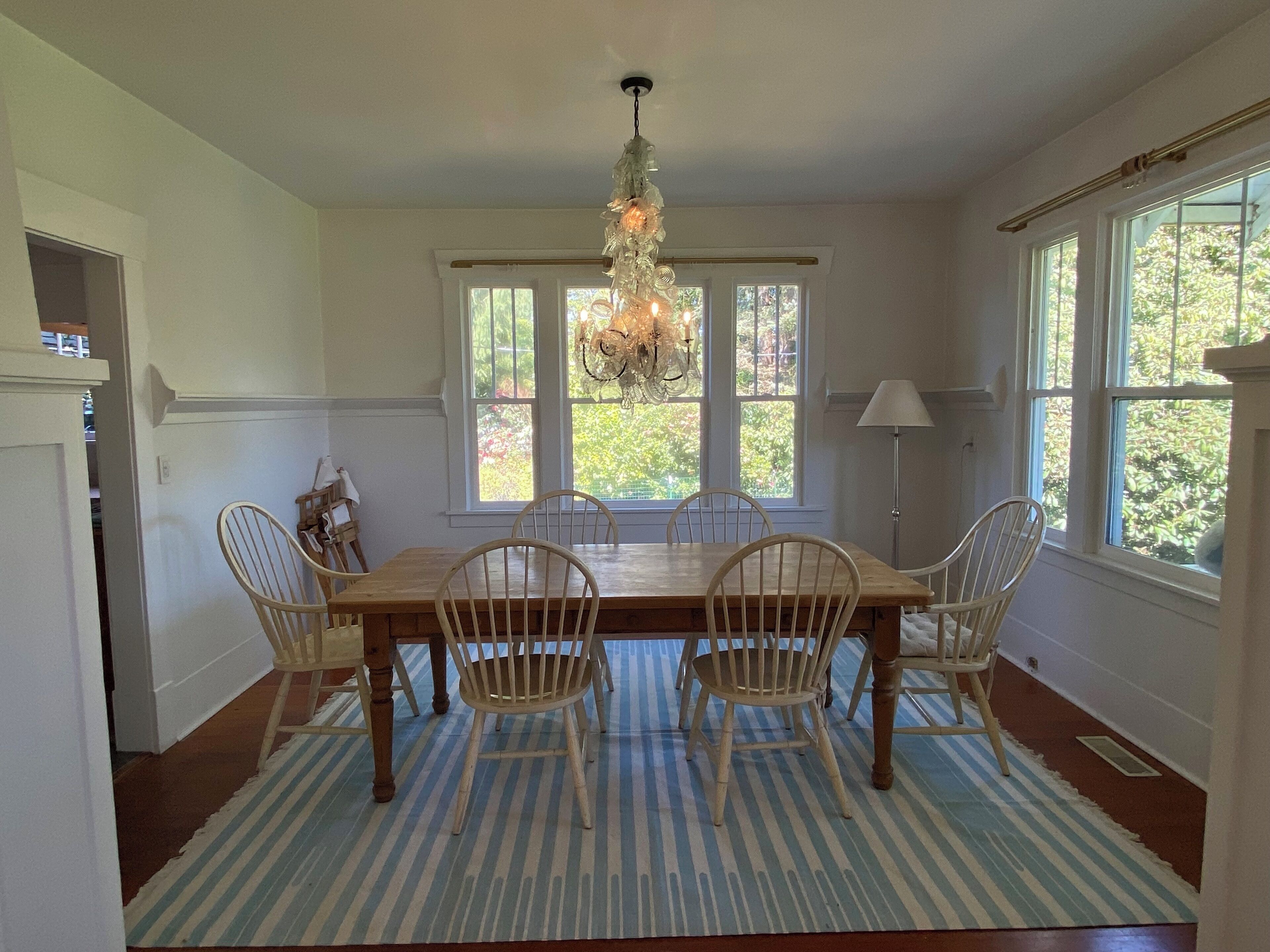 A dining room with a wooden table, four chairs, a chandelier, large windows, and a striped rug.