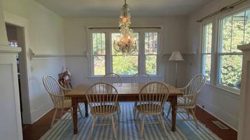 A dining room with a wooden table, four chairs, a chandelier, large windows, and a striped rug.
