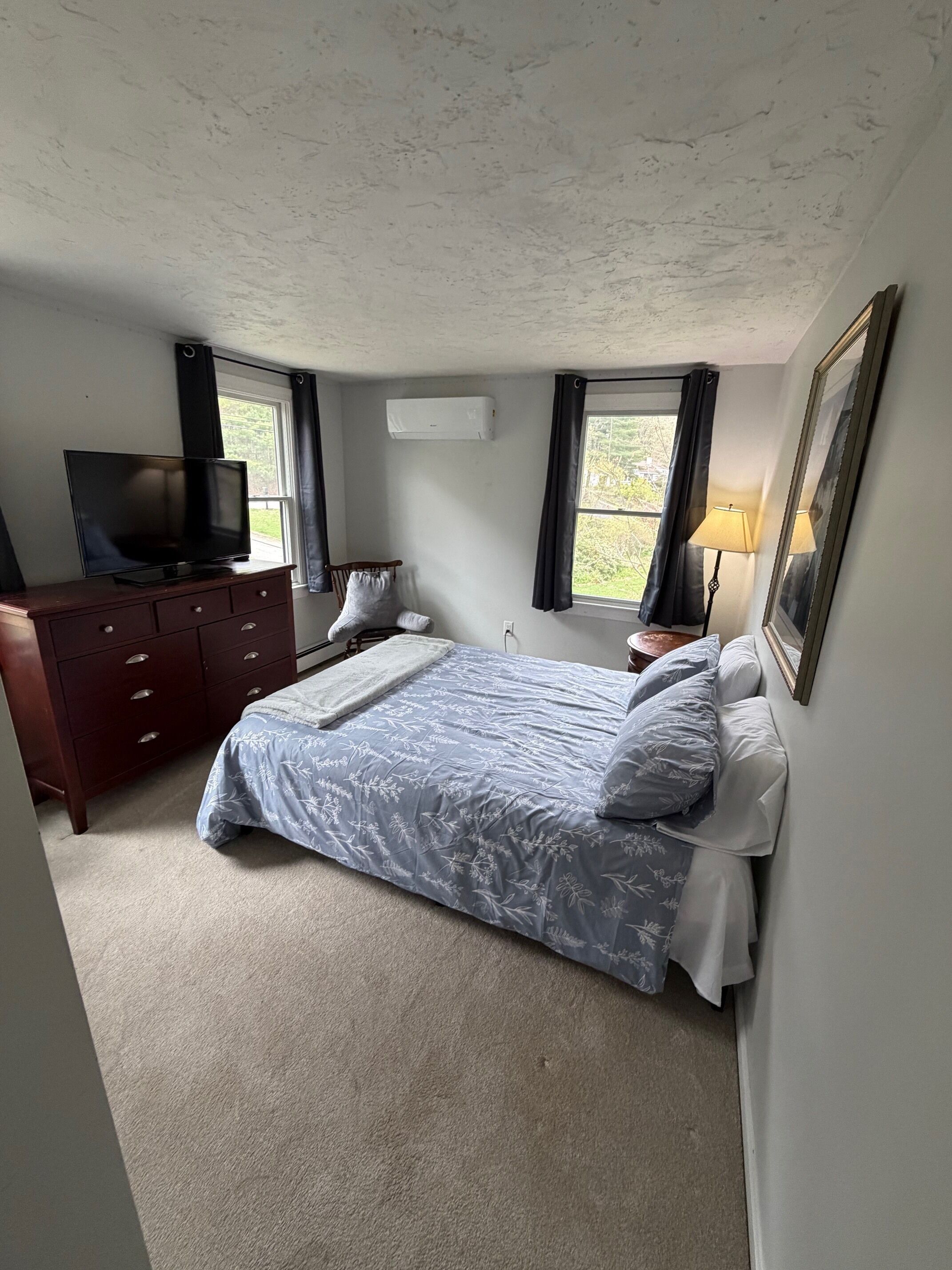 Ein Schlafzimmer mit Bett, Kommode, Fernseher, Fenstern und einer Lampe.