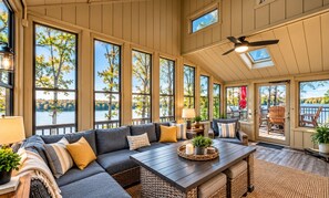 A spacious room with a large sofa, a dining table, and multiple windows offering views of a lake and trees.