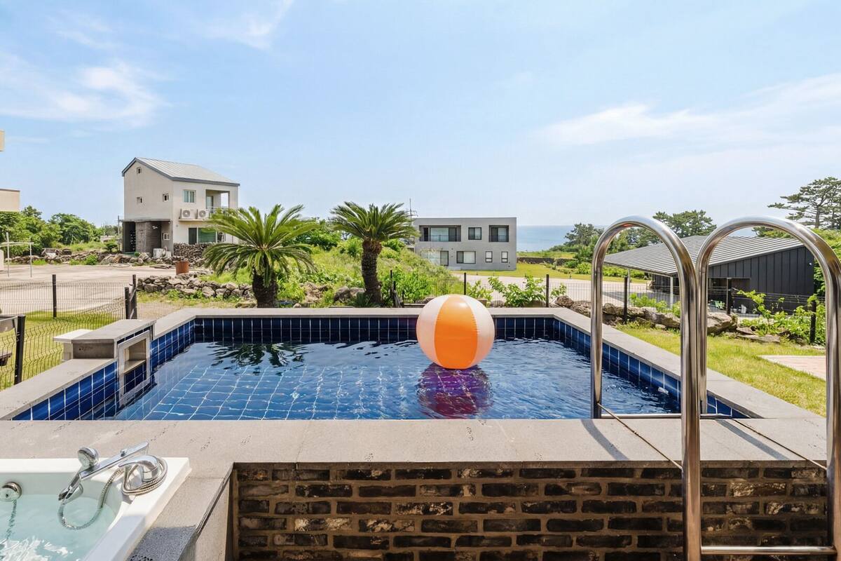 A swimming pool with a beach ball, a hot tub, and a view of buildings and greenery.