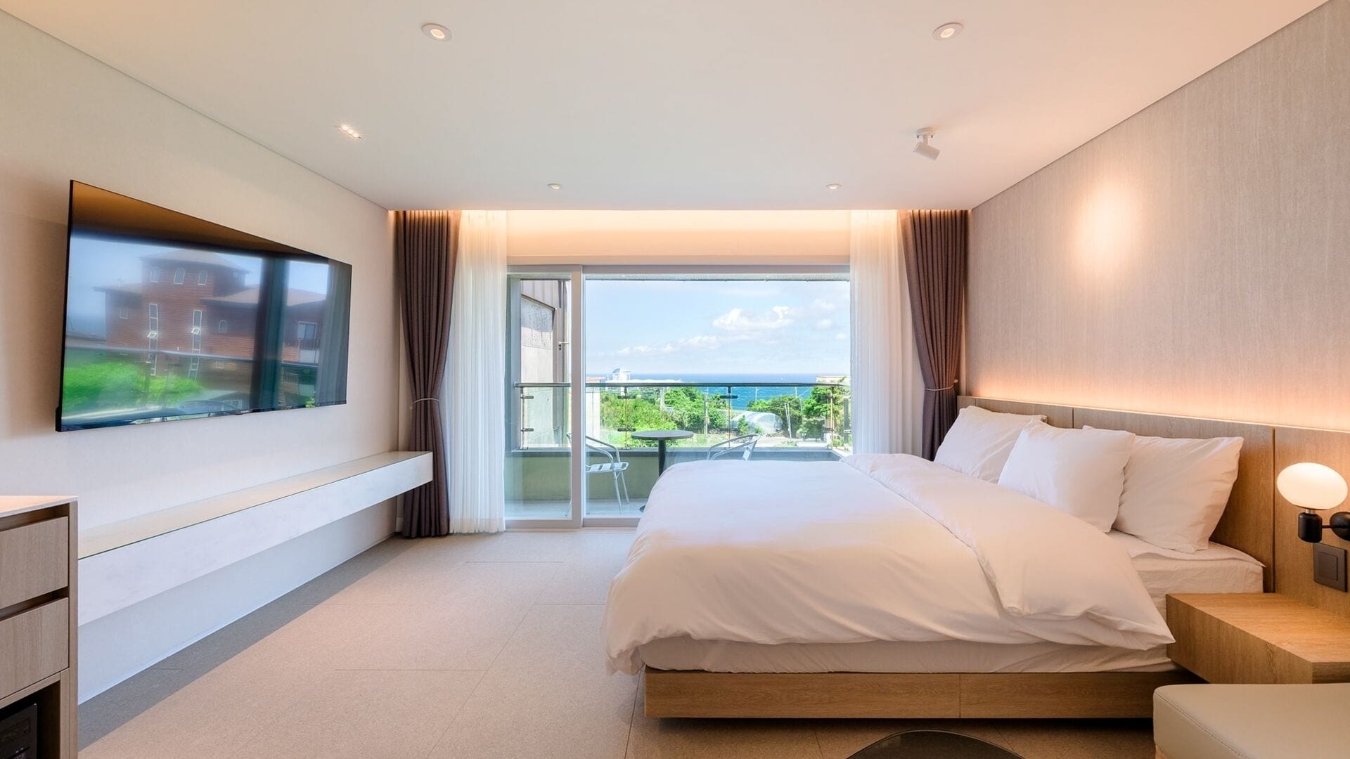 Una habitación de hotel moderna con una cama grande, un televisor montado en la pared y un balcón con vistas al mar.