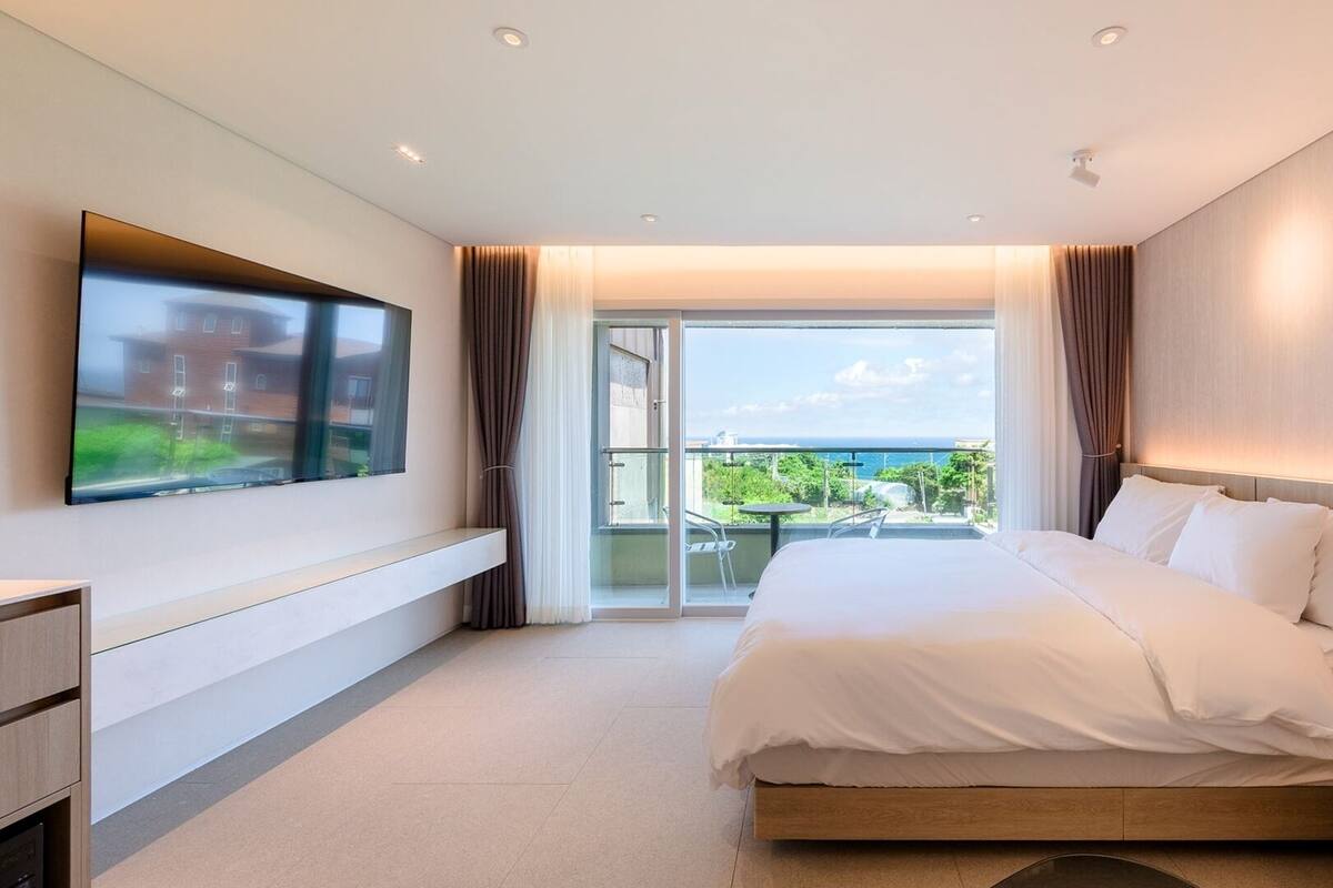 A modern hotel room with a large bed, a TV mounted on the wall, and a balcony with a view of the sea.