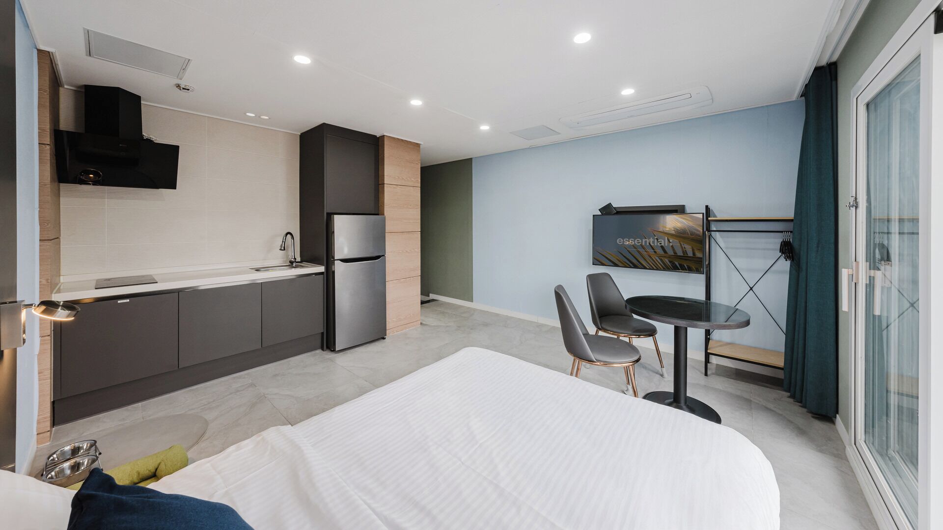 Una habitación de hotel moderna con cama, kitchenette, zona de comedor y un ventanal grande.