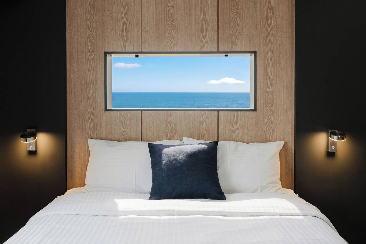 A bedroom with a bed, a window with a view of the sea, and wall-mounted lights.