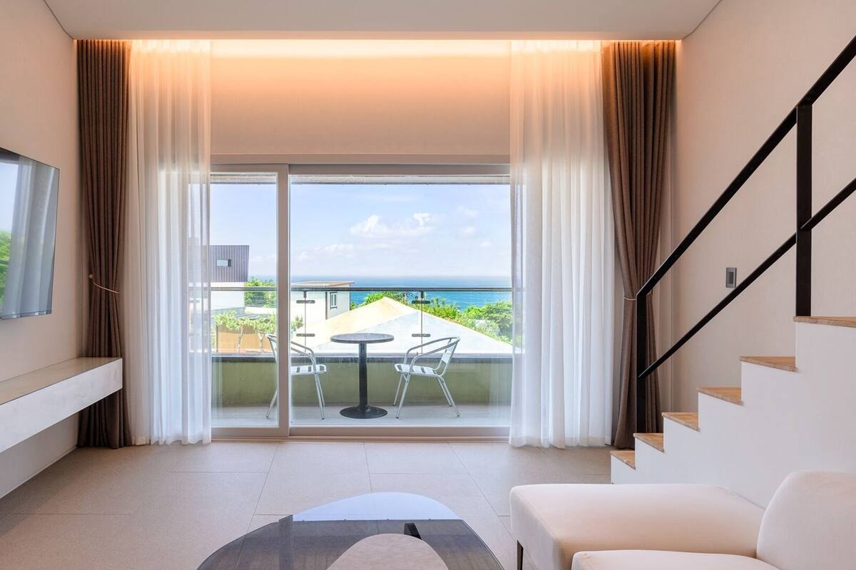 A modern hotel room with a balcony, a flat-screen TV, a sofa, and a staircase.