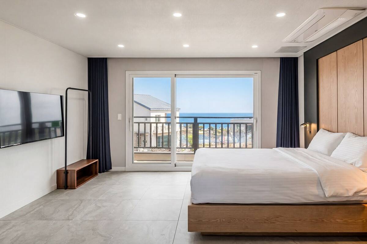 A modern hotel room with a large bed, a flat-screen TV, a balcony with a view of the sea, and a sliding glass door.