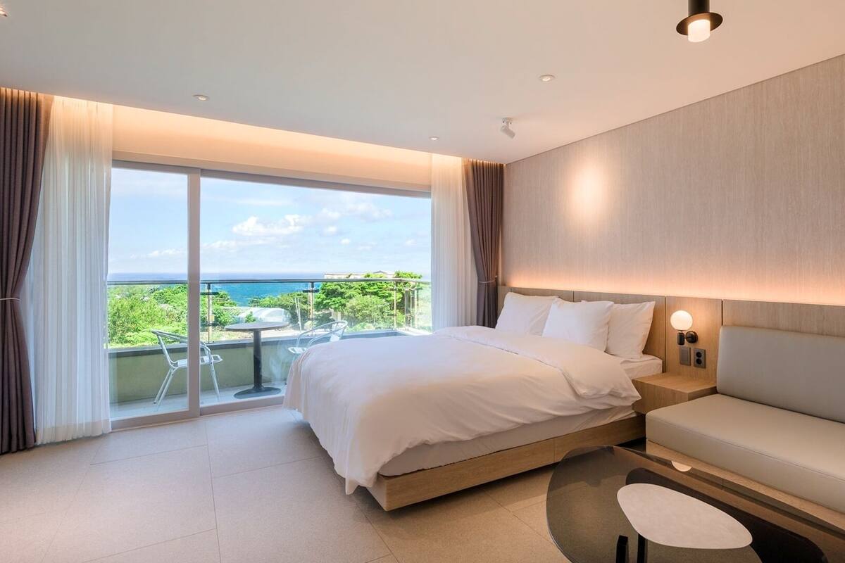 A modern hotel room with a large bed, a sofa, a small table, and a balcony with a view of the sea.