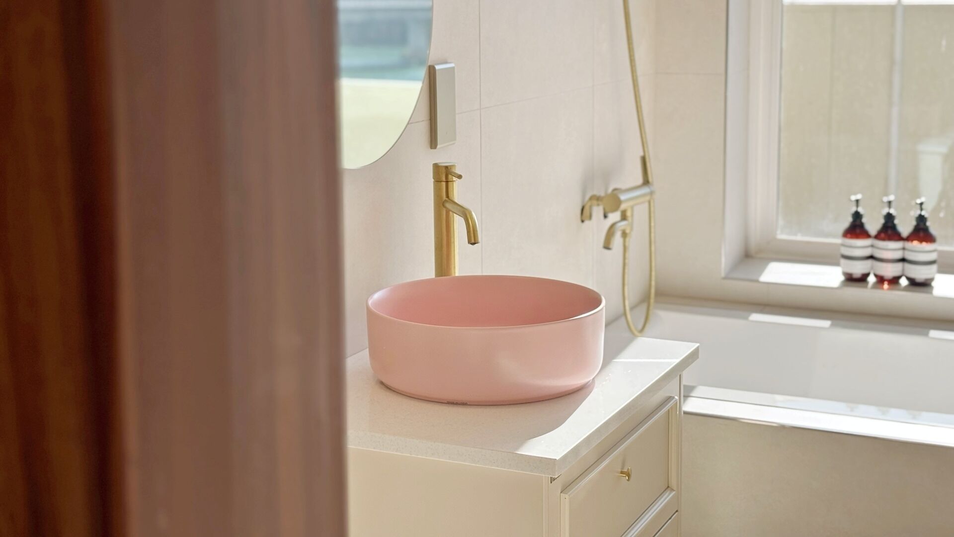 A bathroom with a pink basin, a gold faucet, and a window with bottles on the sill.