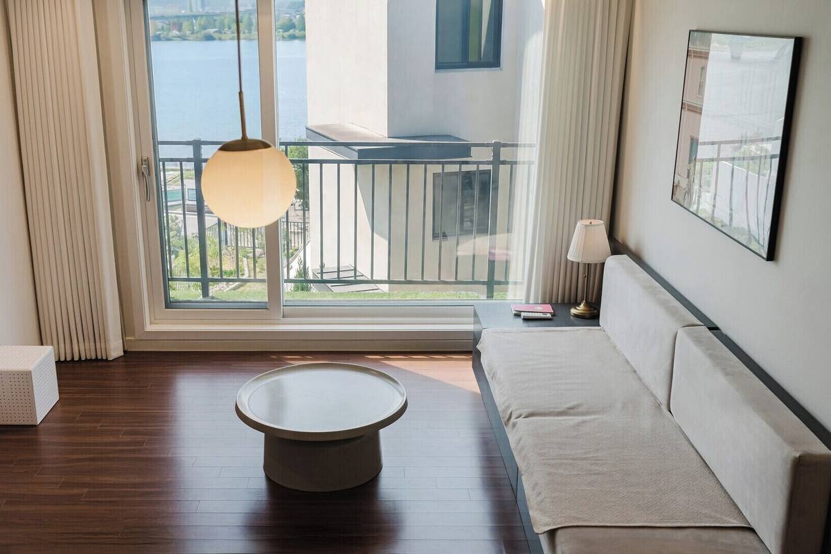 Ein modernes Zimmer mit einem runden Tisch, einer Couch, einem Balkon mit Geländer und Blick auf ein Gewässer.
