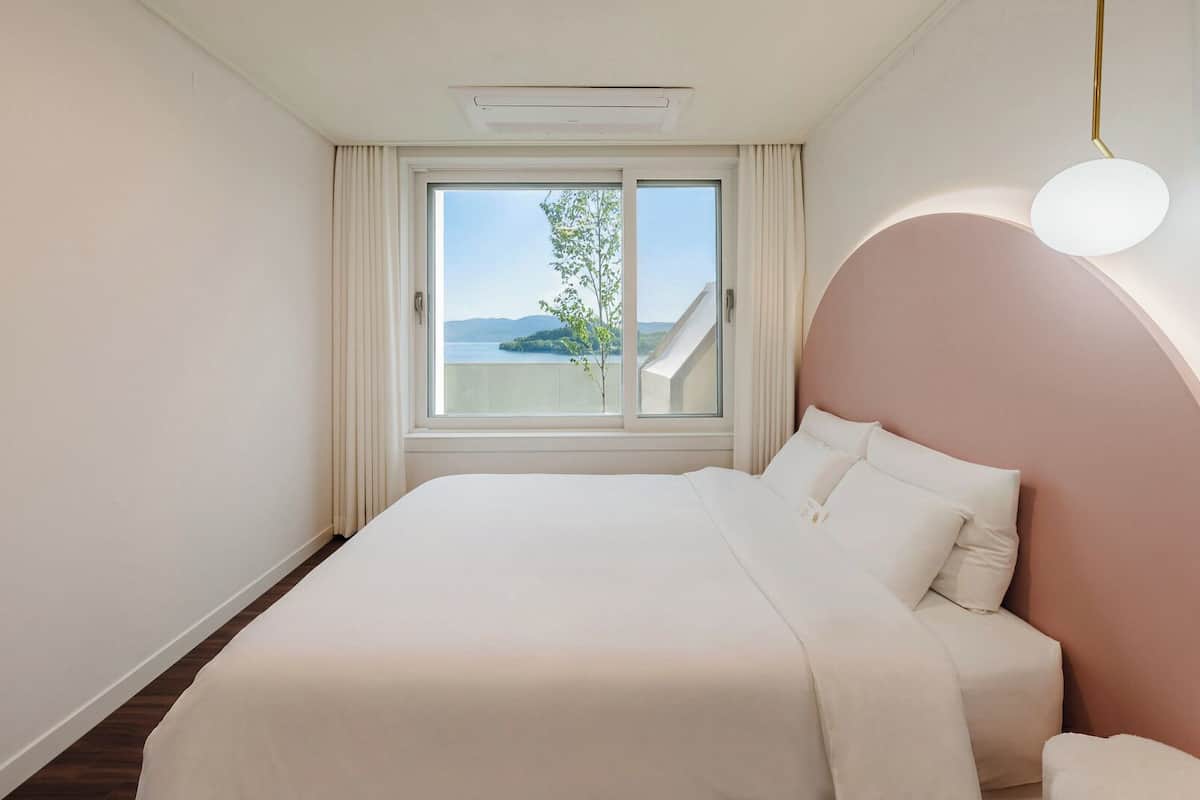 Ein Hotelzimmer mit Bett, einem fenster mit Vorhängen, Blick ins Freie und einer einzigartigen Deckenleuchte.