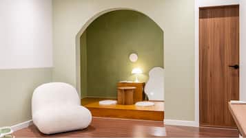 A room with a white chair, a small wooden table with a lamp, and a door.