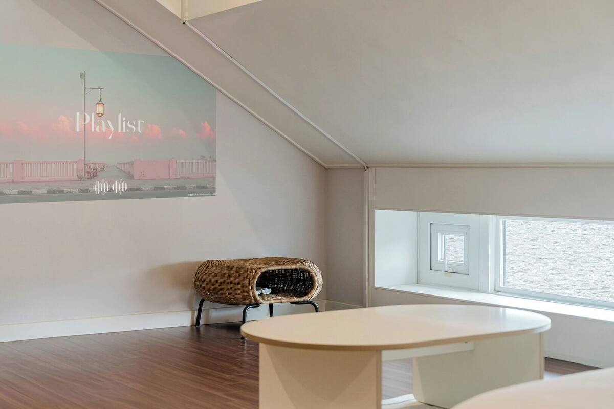 Ein modernes Zimmer mit schrägen Wänden, einem großen Fenster, einem Korbstuhl, einem weißen Tisch und einem an der Wand montierten Projektor, der eine 'Playlist' mit einer Straßenszene zeigt.