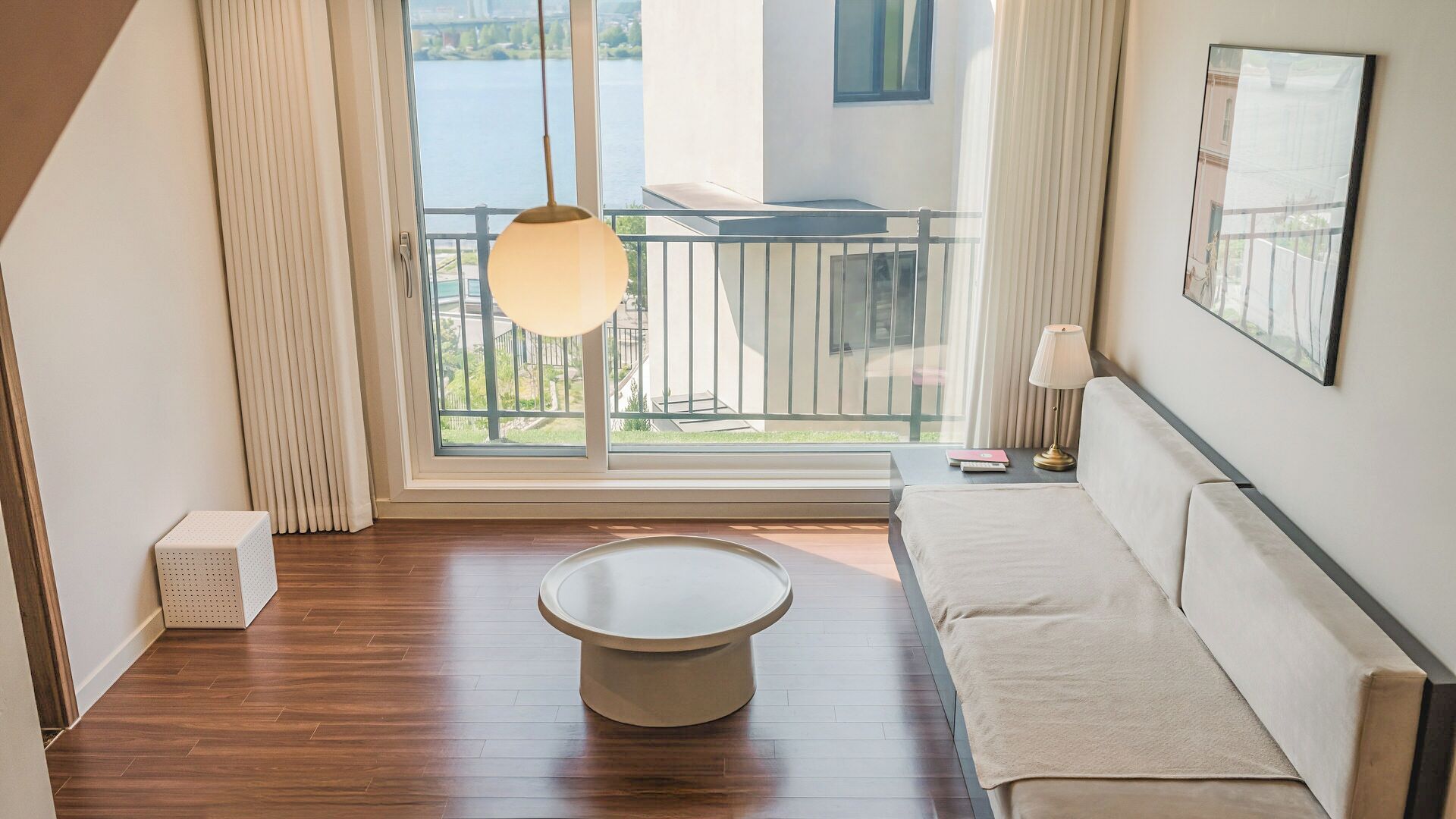 A modern hotel room with a wooden floor, a round coffee table, a sofa, a balcony with a view of the water, and a hanging lamp.